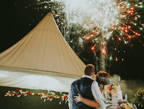 Against a backdrop of red and green fireworks lighting up the night sky, a couple stands embraced, their arms wrapped around each other's backs. In the background, a stunning wedding tipi stands tall, blending seamlessly with the natural beauty of the surrounding environment. The vibrant colours of the fireworks reflect off their faces, filling them with wonder and joy. This moment is pure magic, an unforgettable experience that speaks to the beauty of nature, the power of love, and the exquisite design of Origami Marquees. Let us help you create your own magical memories with our tipi hire services, the perfect addition to any special occasion.