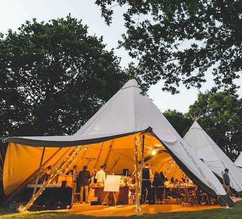 White Tipi hire for a wedding at dusk with orange glowing fairy lights wrapped around the wooden pillars.