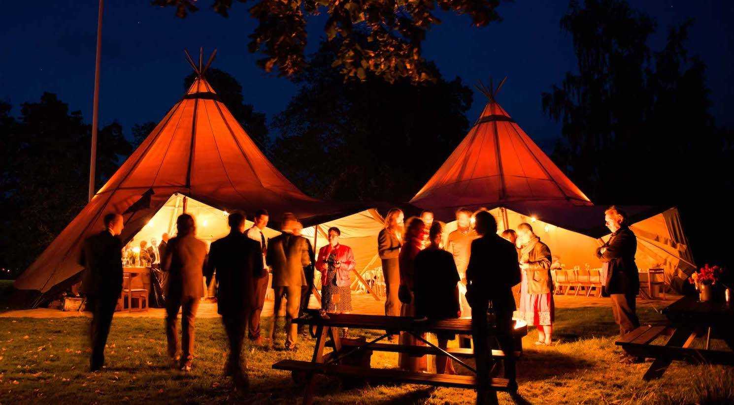 A tipi set up for a corporate event at night, illuminated by strings of lights and with elegant furniture inside. The tipi is located in a natural setting, surrounded by trees and greenery, and the starry night sky can be seen overhead.