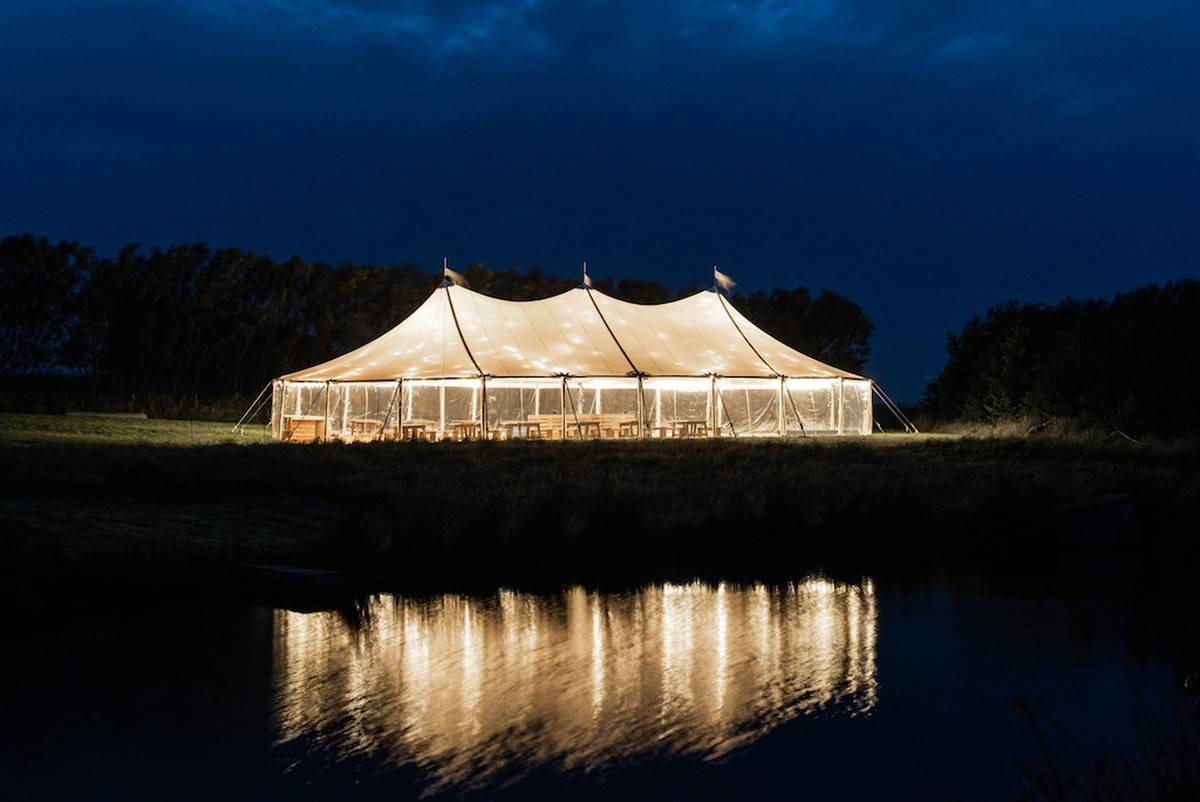 A festival Sailcloth tent, designed for a corporate event, is reflected in a serene lake surrounded by lush greenery, offering a harmonious and captivating setting.