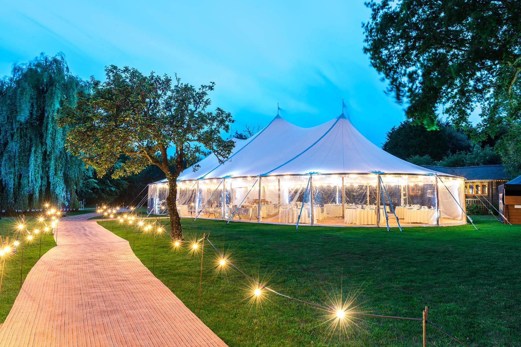 The exterior of a Sperry Tent at night with a festoon lighting walkway leading up to it.