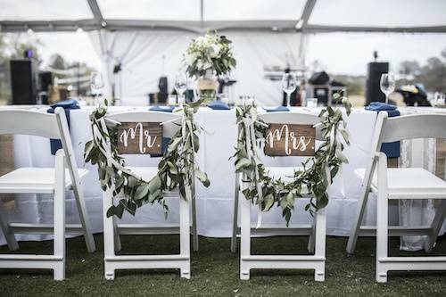 A beautifully arranged wedding reception table beneath a marquee, adorned with charming Mr and Mrs signs on the backrests of the chairs, all provided by Birmingham Marquee Hire.